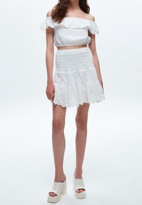 Femme portant un haut court blanc à épaules dénudées et une mini-jupe à volants assortie avec des sandales à talons compensés, debout devant un fond blanc.