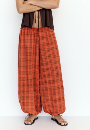 Personne portant un pantalon large ample à carreaux orange avec liens aux chevilles et des sandales plates, debout les bras croisés devant un fond blanc.