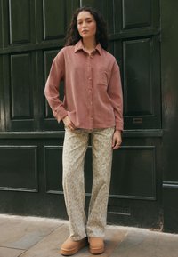 Camicia di velluto a coste color ruggine con bottoni frontali e tasca sul petto, abbinata a pantaloni beige con stampa leopardata e scarpe con platform color cuoio.