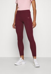 Leggings burdeos con un diseño ajustado, hechos de material elástico, con un pequeño logo en la cadera, combinados con zapatillas deportivas blancas.