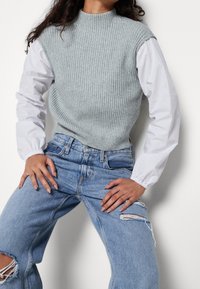 Maglione smanicato a coste grigio chiaro sovrapposto a una camicia bianca a maniche lunghe, abbinato a jeans blu strappati, che mostrano un fit rilassato e texture interessanti.
