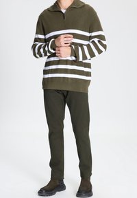 Hombre vestido con un suéter a rayas verde oliva y blanco, pantalones verde oliva oscuros y botas de ante verde oscuro, de pie frente a un fondo sencillo.