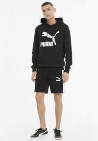 Svart hettegenser med hvit Puma-logo, sammen med svarte shorts med sidestrekk, og svarte sneakers. En enkel, atletisk design.