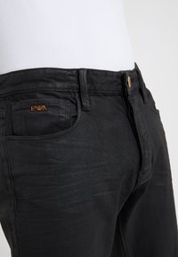 Černé džíny z denimu s  úzkým střihem, zdůrazňující hladkou texturu, hnědá štítek s logem na přední kapse a kovové zapínání na knoflík.