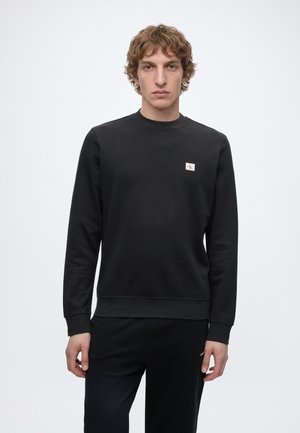 Młody mężczyzna o kręconych włosach, ubrany w jednolitą czarną bluzę z małym, białym i czerwonym logo Calvin Klein na piersi na białym tle.