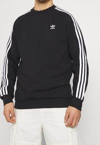 Svart sweatshirt i bomull, med vita tre-randsdetaljer på ärmarna och en liten vit Adidas-logotyp på bröstet.