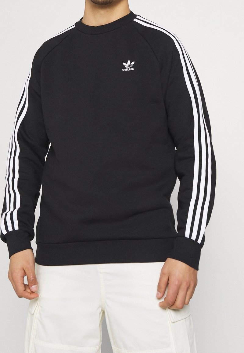 Svart sweatshirt i bomull, med vita tre-randsdetaljer på ärmarna och en liten vit Adidas-logotyp på bröstet.