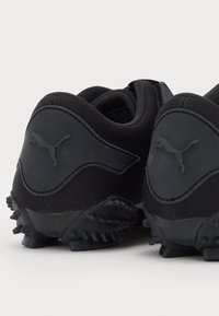 Zapatillas deportivas negras con una parte superior de malla texturizada y acentos de goma, que presentan un logotipo destacado en el talón y una suela con clavos para mayor tracción.