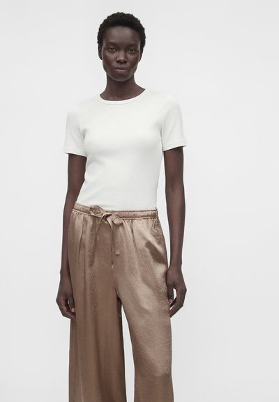 Γυναίκα με κοντά σγουρά μαλλιά, που φοράει λευκό κοντομάνικο πουκάμισο και φαρδιά, γυαλιστερά μπεζ παντελόνια με δεμένη μέση, στέκεται μπροστά σε ένα μονόχρωμο φόντο.