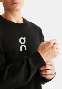 Black sweatshirt with a white logo on the chest, featuring a crew neckline and ribbed cuffs. Fabric appears soft and textured.