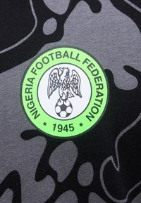 Λογότυπο της Ποδοσφαιρικής Ομοσπονδίας της Νιγηρίας με έναν αετό που στέκεται πάνω σε μια ποδοσφαιρική μπάλα, φωτεινό πράσινο κύκλο και το "1945" σε σκουρόχρωμο φόντο παραλλαγής υφάσματος.