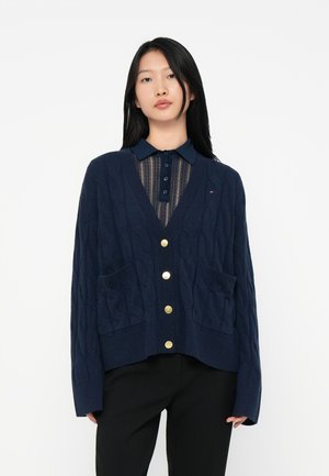 Young woman with straight black hair wearing a navy blue buttoned cable knit cardigan over a sheer striped navy top and black pants.