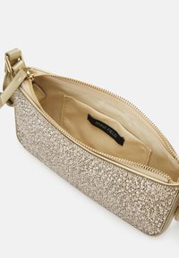 Petit sac à main doré avec extérieur pailleté, intérieur beige, fermeture éclair sur le dessus et une poche intérieure avec une étiquette noire "Anna Field".