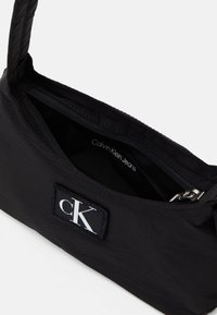 Borsa di tessuto nero con una texture liscia, caratterizzata da una toppa con il logo e un'apertura con cerniera. Interno con etichetta "Calvin Klein Jeans".