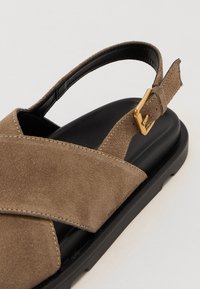 Beez-suede sandaalid beežis, ristiribade disain, must kummist tald ja reguleeritav rihm, millel on kuldne tasku. Sile tekstuur ja minimalistlik välimus.