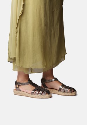 Ben iført bronze metaliske lukkede flet-sandaler med ankelremme og flettet jutesål, under en flydende olivengrøn plisseret kjole.