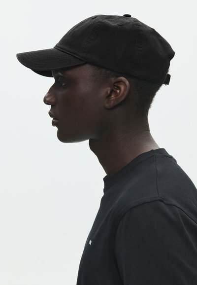 Fiatal fekete férfi fekete baseballsapkában és fekete ingben, balra néz, egyszínű világos háttér előtt.