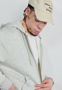 Lysegrå zip-up hoodie med en glat tekstur og forlommer, båret over en hvid t-shirt. Begyndt med en beige kasket med broderet tekst.
