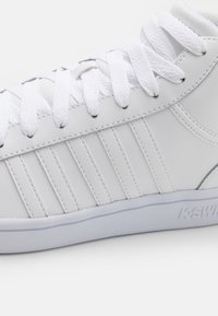Zapatilla de piel blanca con cordones blancos, rayas verticales cosidas en el costado y "K-Swiss" grabado en la suela.