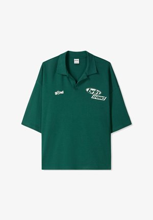 Zelena kratka polo majica ohlapnega kroja, z ovratnikom in belimi napisi ter logotipi na sprednji strani.