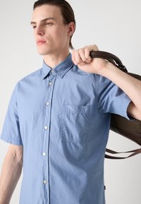 Lichtblauwe shirt met korte mouwen, knoopsluiting aan de voorkant, spitse kraag en een borstzakje. De stof heeft een licht gekreukt textuur.