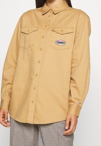 Camicia button-up marrone chiaro con due tasche sul petto e una patch con il logo "TOMMY", realizzata in tessuto testurizzato e con colletto.