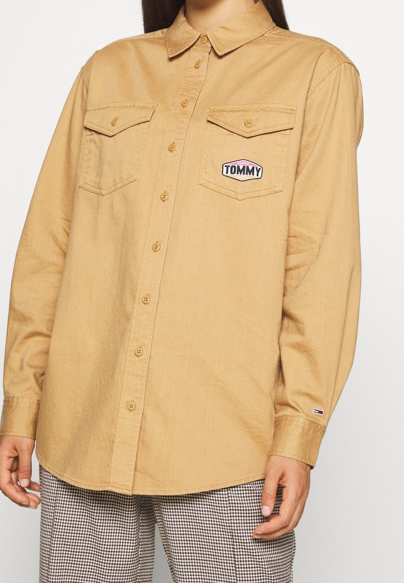 Camicia button-up marrone chiaro con due tasche sul petto e una patch con il logo "TOMMY", realizzata in tessuto testurizzato e con colletto.