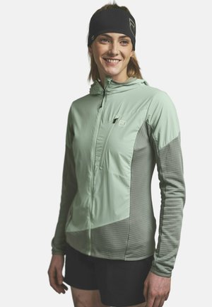 Lächelnde Frau, die eine hellgrüne Jacke mit Reißverschluss trägt, die strukturierte graue Ärmel hat, und ein schwarzes Stirnband, steht vor einem schlichten weißen Hintergrund.