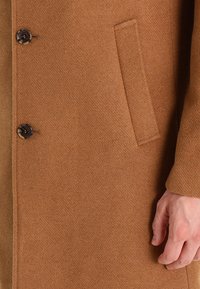 Brun kappa i ullblandning med en texturerad yta, två knappar och en sidoficka. Designen är skräddarsydd med en slät finish.