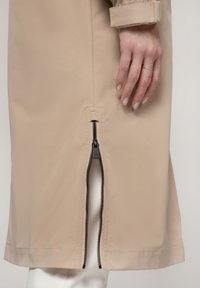 Beige regenjas met een zijrits en een zwarte metalen trekker, van textuurstof en met een rechte zoom. Handmodel toont de details van het kledingstuk.