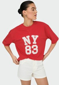 Rode oversized t-shirt met witte opdruk "NY 83". Korte mouwen, ronde halslijn, gecombineerd met witte denim shorts. Gladde katoenstructuur.