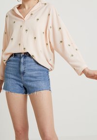 Kobieta w jasnopomarańczowej bluzce w niewielkie nadruki palm, w wysokich niebieskich dżinsowych szortach z postrzępionymi nogawkami.