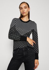 Černý svetr s šedobílým chevron vzorem, s texturovaným povrchem a černými žebrovanými manžetami a lemem. V kombinaci s tmavými džíny.