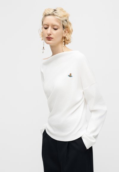 Blond osoba v bielom dlhom tričku s malým farebným logom planéty, čiernych nohaviciach a s dlhými visiace náušnicami, na jednoduchom pozadí.