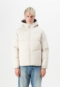 Chaqueta puffer de color beige claro con capucha, con acolchado de franjas horizontales, cremallera frontal y puños elásticos, llevada sobre jeans de mezclilla azules.