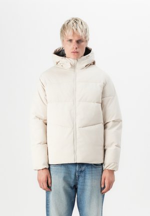 Chaqueta puffer de color beige claro con capucha, con acolchado de franjas horizontales, cremallera frontal y puños elásticos, llevada sobre jeans de mezclilla azules.