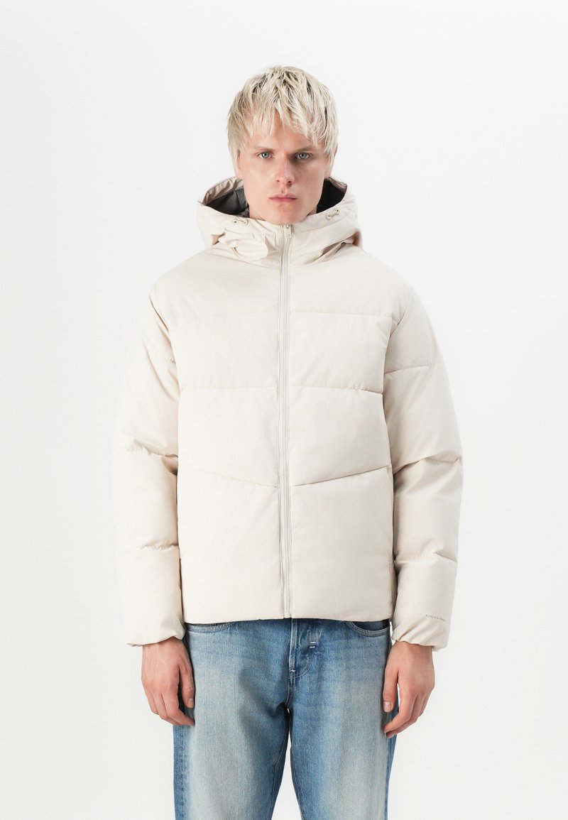 Chaqueta puffer de color beige claro con capucha, con acolchado de franjas horizontales, cremallera frontal y puños elásticos, llevada sobre jeans de mezclilla azules.