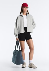 Frau mit roter Mütze, grauer Reißverschlussjacke, weißem Hemd, schwarzer Shorts, weißen Socken, Turnschuhen, die eine große blaue Tragetasche hält.