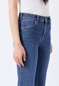 Jeans de tiro alto de color azul hechos de mezclilla con una textura suave, que cuentan con cinco bolsillos, remaches de cobre y sutiles detalles de costura.