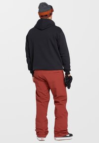 Zwarte hoodie, roestkleurige broek en zwarte handschoenen. Grijze beanie met rode accenten. Sneakers met witte zolen en bruine details.
