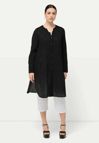 Schwarze lange Tunika mit dezentem Strukturmuster, runder Halsauschnitt, Knopfverschluss vorne und seitlichen Schlitzen, kombiniert mit kurzen weißen Hosen und schwarzen hohen Schuhen.