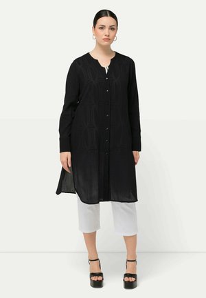 Schwarze lange Tunika mit dezentem Strukturmuster, runder Halsauschnitt, Knopfverschluss vorne und seitlichen Schlitzen, kombiniert mit kurzen weißen Hosen und schwarzen hohen Schuhen.