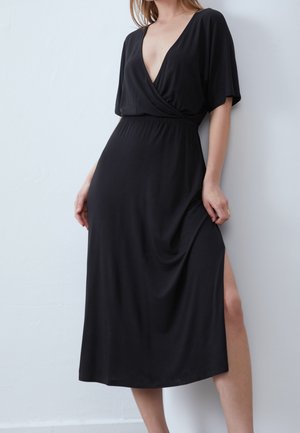 Femme portant une robe midi noire avec un décolleté en V profond, des manches courtes, une taille cintrée et une fente latérale haute jusqu'à la cuisse, debout contre un mur blanc.
