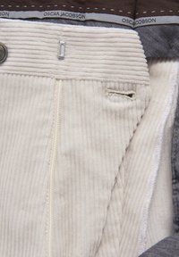 Beige manchestertyg med vertikala ränder, utrustad med en dragkedja och en ögla för justering. Fodrad med grått tyg med synliga söm detaljer.