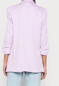 Blazer lilas clair avec une coupe ajustée, manches trois-quarts, et une texture de tissu lisse. Comprend une fente unique au dos et sans fermetures visibles.