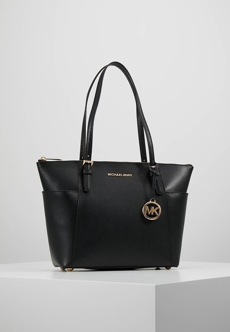 Sac cabas en cuir noir Michael Kors avec doubles poignées, breloque logo dorée et poches latérales, présenté sur une surface blanche.