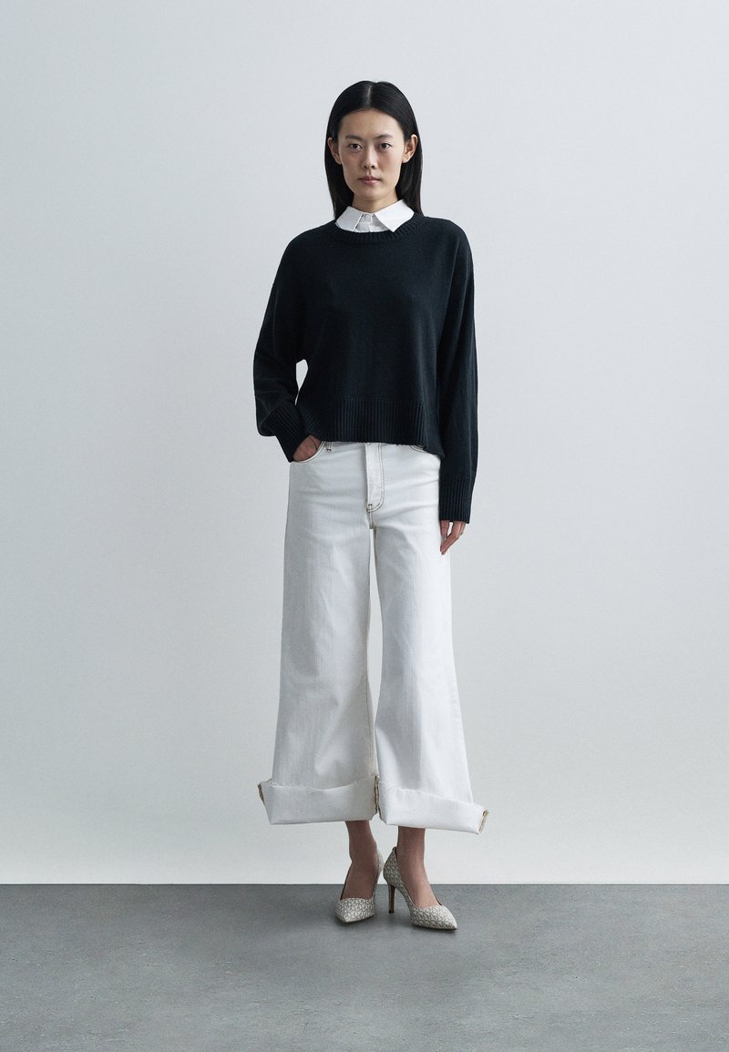 Maglione nero in maglia con orlo a coste, abbinato a jeans bianchi a gamba larga con risvolti. Calzature: scarpe con tacco alto e texture.