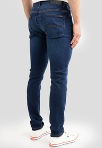 Muž v tmavě modrých slim fit džínách a bílých teniskách, zobrazený zezadu na jednobarevném světlém pozadí.