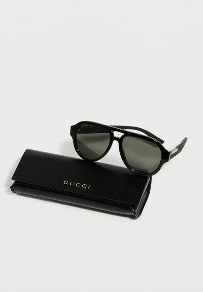 Μαύρα γυαλιά ηλίου Gucci με σκοτεινούς φακούς, ακουμπισμένα πάνω σε μαύρη θήκη με το λογότυπο Gucci, πάνω σε λευκό φόντο.