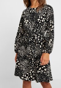 Černé a krémové šaty se vzorem, dlouhými rukávy, páskem v pase a vrstevnatým volánkovým lemem. Obsahují abstraktní tvary a pohodlný siluetu.